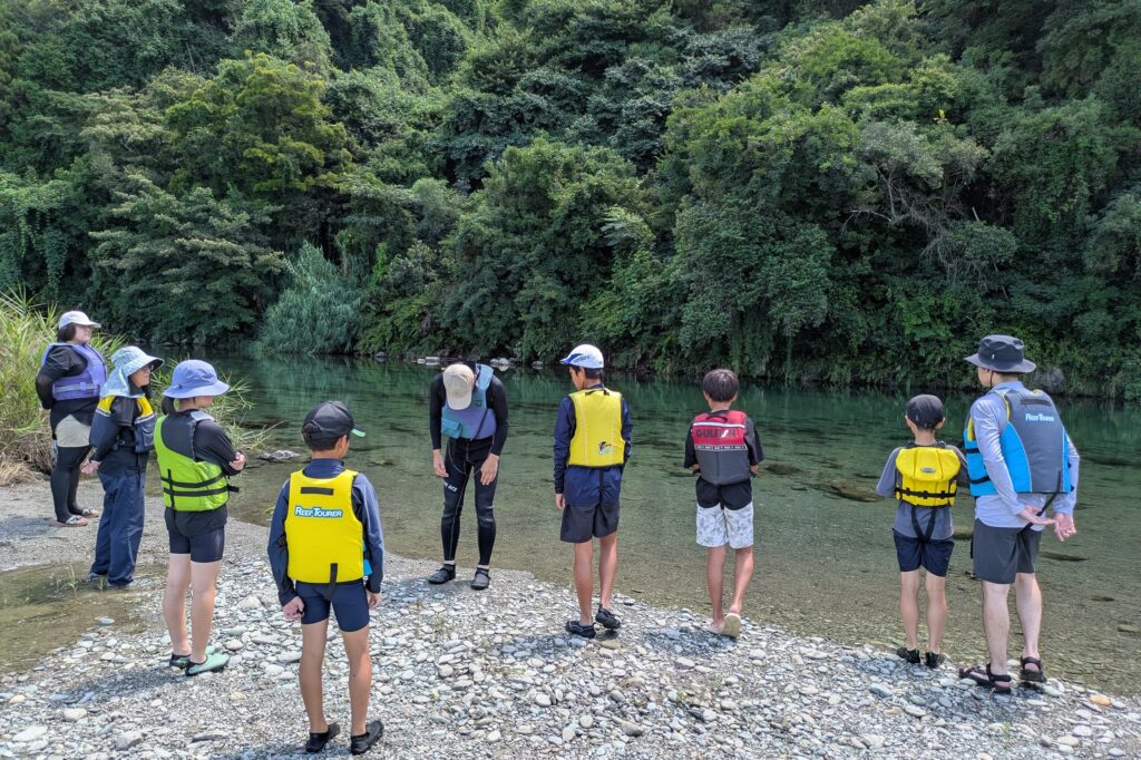 川の深さはどのくらいだと思う？、と子どもたちに問いかける西条自然学校の山本貴仁さん（中央）